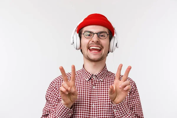 Musique, technologie et concept humain. Jeune homme souriant positif et heureux avec barbe et lunettes, porter un bonnet écouter de la musique dans les écouteurs, montrant un geste de paix et de rire, fond blanc — Photo