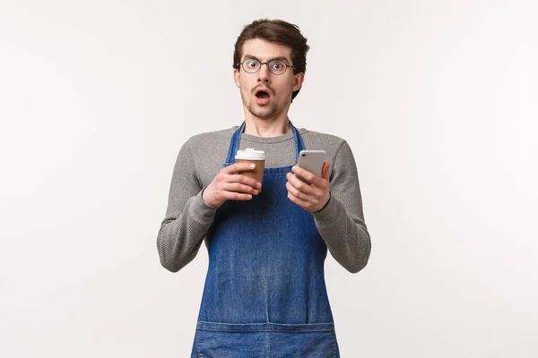 Porträtt av chockad kille i dumhet, bär förkläde som arbetar i kafé eller bardisk, hålla mobiltelefon och kaffe i take-away cup kippar och stirra chockad kamera, läsa skakade nyheter — Stockfoto