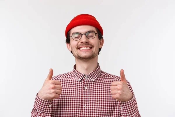 ¡Muy bien! Primer plano retrato satisfecho guapo joven con bigote, usar gorro rojo y gafas, cerrar los ojos y sonreír complacido, mostrar los pulgares hacia arriba como cosa totalmente impresionante, fondo blanco — Foto de Stock