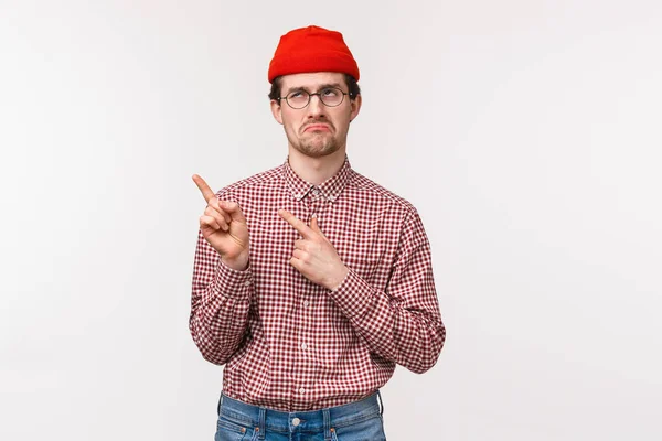 Skeptisk och oroad uttråkad ung hipster kille i röd mössa och glasögon, grimma irriterad på att besöka tråkigt möte, peka övre vänstra hörnet, ögonrulla och sura störd — Stockfoto