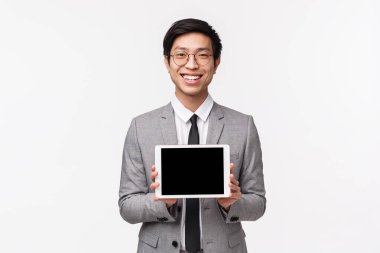 Gri takım elbiseli ve gözlüklü hoş görünümlü Asyalı adamın belden yukarı portresi, başvuru, proje veya finansal baskı tanıtımı, dijital tablet ekranı gösterme ve tatmin olmuş gülümseme