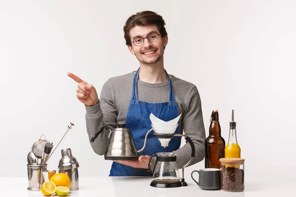 Barista, trabajador de cafetería y camarero concepto. Retrato de encantador empleado masculino sonriente, dueño de la tienda sosteniendo hervidor de agua y señalando el dedo a la izquierda, preparar café de filtro, de pie fondo blanco — Foto de Stock