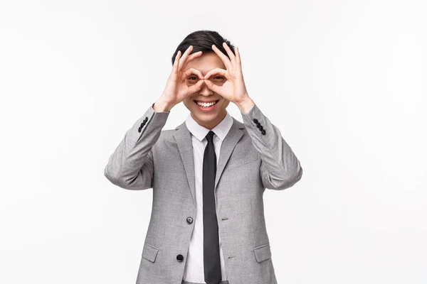 Waist-up portrait of funny and cute, playful young asian male in suit, entrepreneur fool around, making finger mask-glasses and looking through it, smiling, seeing something interesting — Stock Photo, Image