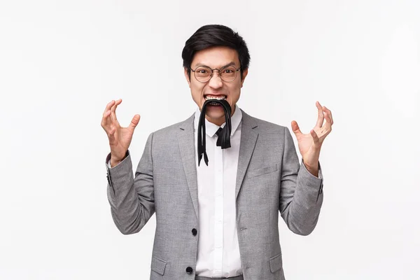 Waist-up portrait of pissed-off, distressed and irritated young asian man eating his own tie and clenching fists aggressive, feeling fed up and annoyed, grimacing furious, white background