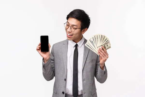 Cintura de afortunado y exitoso joven hombre de negocios de TI en traje gris, enriqueciéndose usando la aplicación, mostrando algo en la pantalla móvil y sosteniendo dólares, gran suma de dinero, fondo blanco — Foto de Stock