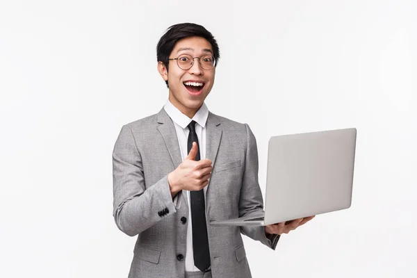 Waist-up portrait of enthusiastic happy young asian man in grey suit, reacting to amazing news or special price offer, using laptop, show thumbs-up and smiling at camera, white background