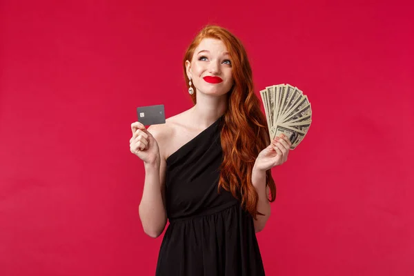 Luxury, beauty and money concept. Dreamy rich and lucky good-looking redhead woman thinking how waste her prize cash, smirk indecisive and looking up, put money on deposit credit card or shopping