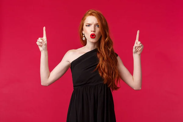 Celebración, eventos, concepto de moda. Retrato de mujer pelirroja frustrada y desconcertada en lápiz labial rojo, vestido elegante negro, frunciendo el ceño decepcionado, señalando algo malo — Foto de Stock