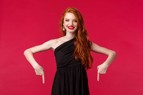 Celebración, eventos, concepto de moda. Retrato de alegre, sonriente joven pelirroja elegante en vestido negro, señalando con los dedos hacia abajo y mirando la cámara feliz, consejo haga clic en el enlace, fondo rojo —  Fotos de Stock