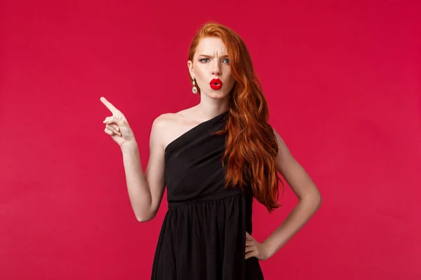 Красота, мода и женская концепция. Разочарованная и сомнительная молодая рыжая женщина, указывающая пальцем влево и хмурящаяся на камеру, расстроена, жалуется или расстроена, носит черное вечернее платье — стоковое фото
