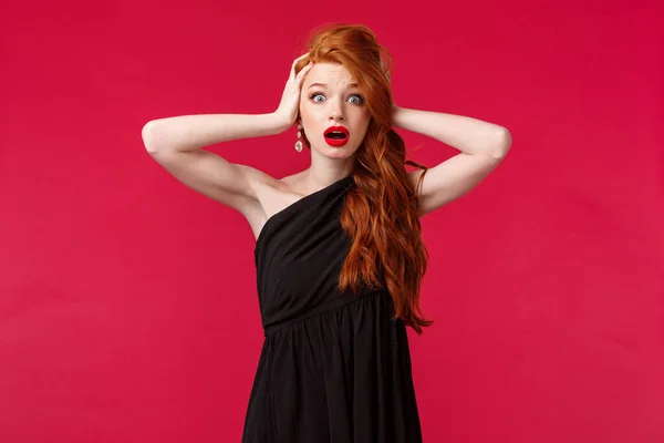 Retrato de mujer pelirroja joven sorprendida y emboscada en elegante vestido negro, jadeando nerviosamente, mirada cámara alarmada cogida de la mano en la cabeza, de pie sobresaltado sobre el fondo rojo —  Fotos de Stock