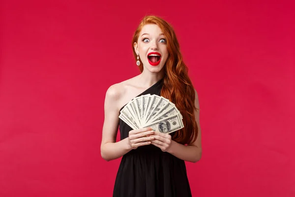 Luxury, beauty and money concept. Surprised and excited gorgeous redhead woman in black dress, screaming amazed and pleased, receive cash prize, stare impressed, feel lucky triumphing