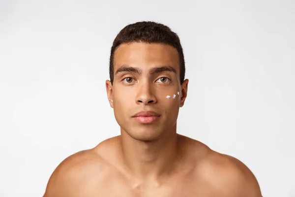 Foto de homem americano africano sem camisa sorrindo e aplicando creme facial isolado sobre fundo branco — Fotografia de Stock