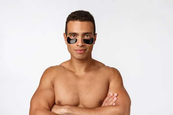 Attractive african young man with under eye black patches caring for his facial skin looking straight at camera on grey background — Stock Photo, Image