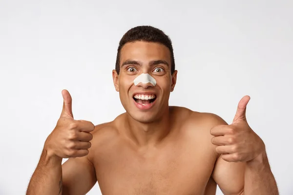 Bonito homem afro-americano usa remendo nasal para reduzir pontos pretos e rugas, usa touca de banho, sorri amplamente . — Fotografia de Stock