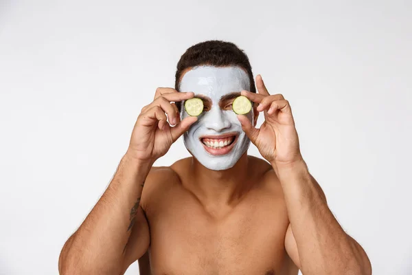 Foto van vrolijke Afrikaanse Amerikaanse man in gezicht cosmetische masker maken plezier met komkommer geïsoleerd over witte achtergrond — Stockfoto