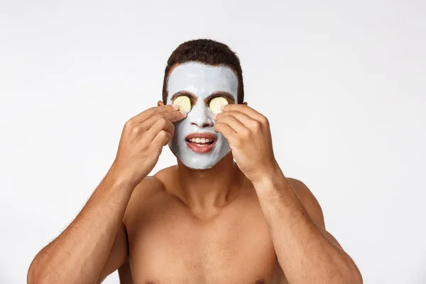 Foto di gioioso uomo afroamericano in maschera cosmetica facciale prendendo in giro con cetriolo isolato su sfondo bianco — Foto Stock