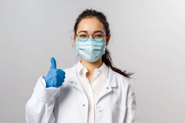 Covid19, koronavirüs, sağlık ve doktor konsepti. İyimser Asyalı kadın doktor portresi her şeyin iyi olacağına dair garanti veriyor. Baş parmak yukarı, virüs kapmamak için tıbbi maske takın. Beyaz önlük.