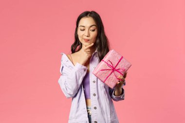 Paketlenmiş kutuya bakan, gülümseyen ve dokunaklı çeneye bakan meraklı Asyalı genç kızın portresi, doğum günü partisinde arkadaşının hediye ettiği pembe arka plan hakkında düşünürken.
