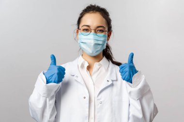 Doktorlar, enfeksiyon uzmanı, araştırma ve kovid19 konsepti. Tatmin olmuş genç Asyalı doktor virüs üzerinde iyi sonuçlar aldı, aşıyı keşfetti, baş parmağını kaldırdı, yüz maskesi ve eldiven taktı.