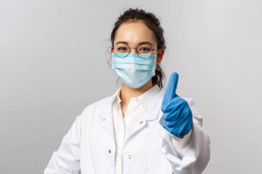 Doktorlar, enfeksiyon uzmanı, araştırma ve kovid19 konsepti. Kendinden emin genç Asyalı kadın doktor başparmağını kaldır, insanları evde kalmaya teşvik et, hastalık virüsüyle savaş, yüz maskesi ve eldiven tak.