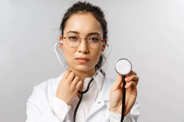 Covid19, coronavirus, gezondheidszorg en artsen concept. Portret van een ernstig uitziende vrouwelijke arts tijdens de controle van de patiënt in ER, ziekenhuis, het dragen van een witte jas, het luisteren naar longen met stethoscoop — Stockfoto