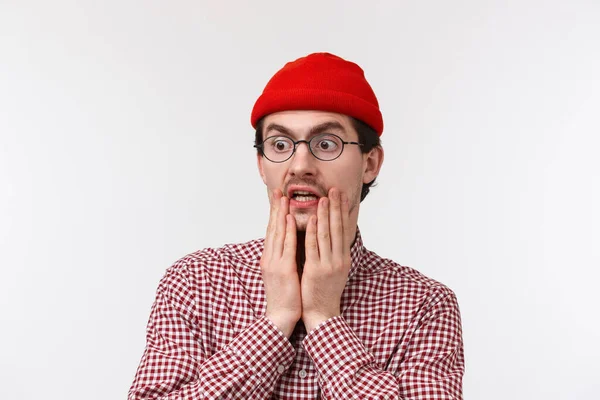Jösses, omg. Förvånad och mållös hipster kille med skägg i röd mössa, glasögon, kippar på något chockerande äckligt, ser vänster plats fruktansvärd sak, stående vit bakgrund — Stockfoto