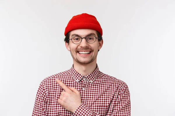 Närbild porträtt glad snygg kaukasiska hipster kille i röd mössa och glasögon, pekar finger övre vänstra hörnet och ler glad, visar reklam, rekommenderar produkt, specialerbjudande — Stockfoto