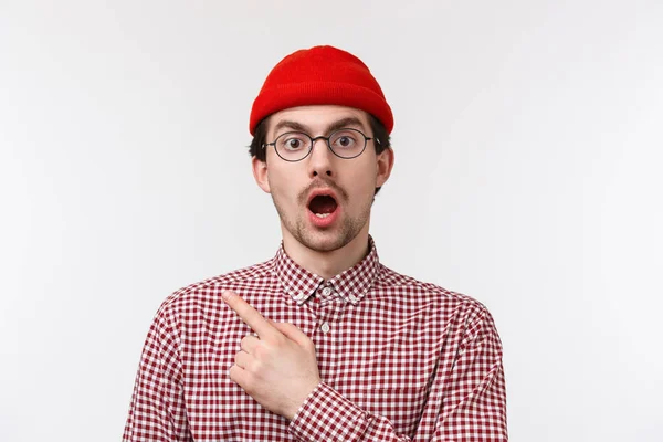 Surpreso e impressionado bonito homem hipster barbudo em óculos e gorro vermelho, boca aberta e mandíbula cair ofegante atordoado, viu algo chocante, apontando canto superior esquerdo, fundo branco — Fotografia de Stock