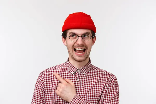 Le gars approuve le choix. Homme caucasien heureux et souriant en bonnet rouge et lunettes pointant vers le coin supérieur gauche, fond blanc . — Photo