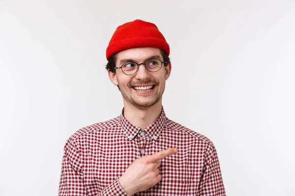 Sly handsome bearded young man in glasses and red beanie, smiling hideous having interesting idea or creative plan, pointing finger upper right corner, standing white background — Stockfoto