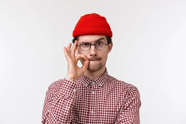 Κοντινό πλάνο πορτρέτο ενός σοβαρού νεαρού Καυκάσου με μουστάκι, φοράει γυαλιά και σκουφί, σφραγίζει τα χείλη με χειρονομίες σαν να κρύβει μυστικό, υπόσχεται να μην μιλήσει γι 'αυτό, στέκεται λευκό φόντο — Φωτογραφία Αρχείου