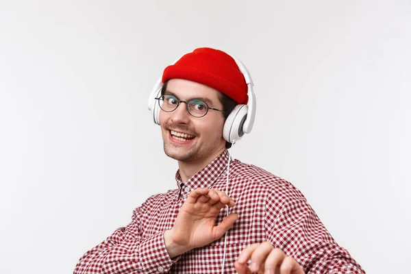 Musica, tecnologia e concetto di persone. Bello sguardo spensierato maschio barbuto in occhiali e berretto rosso ballare in cuffia, fotocamera sorridente godendo playlist cool, stand sfondo bianco — Foto Stock