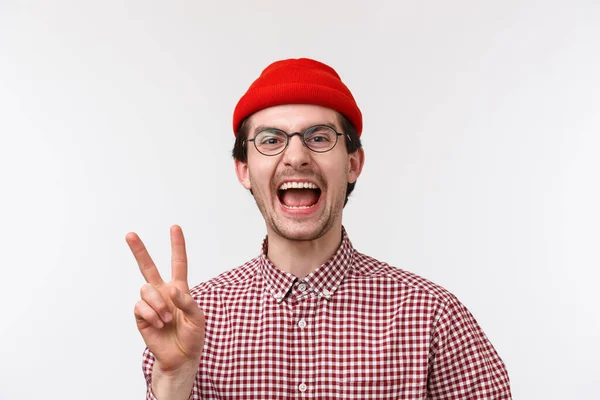 Close-up retrato alegre e engraçado caucasiano barbudo macho em óculos e gorro vermelho, rindo e mostrando gesto de paz olhando câmera feliz, de pé fundo branco — Fotografia de Stock