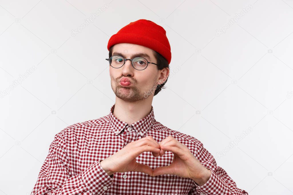 Close-up portrait funny caucasian male with geeky look, wear glasses and red beanie, pouting for kiss, showing heart gesture as confessing love, expressing sympathy in cringy way, white background