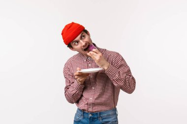 Kutlama, tatiller ve yaşam tarzı konsepti. Sevimli ve kaygısız genç hippi bıyıklı, kırmızı bereli adam, tatlı yerken, tatlı yerken, beyaz arka planda...