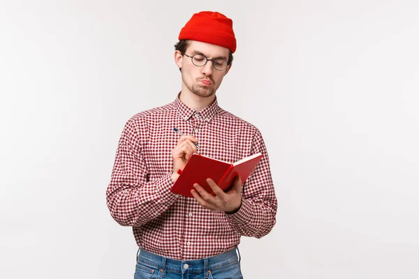 Midja upp porträtt av allvarliga utseende bossig manlig chef i röd mössa, glasögon, sura tankeväckande medan du skriver ner anteckningar i röd anteckningsbok, göra schema eller planera sin dag, vit bakgrund — Stockfoto