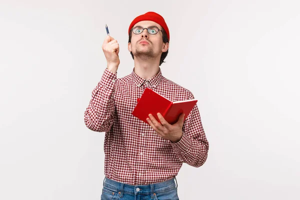 Waist-up portrait of funny male scientist working on project, passionatly look up and raise pen as have perfect idea, holding notebook, writing down his inspiring thoughts, stand white background