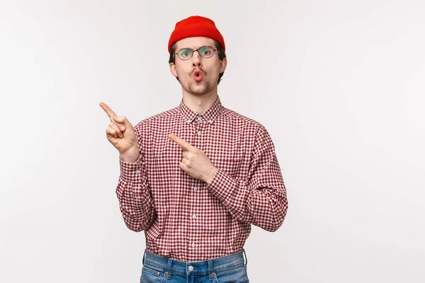 Cintura-up retrato impressionado e intrigado jovem caucasiano barbudo homem em óculos, gorro vermelho, apontando canto superior esquerdo e olhar câmera curiosa, perguntando sobre o produto, fundo branco — Fotografia de Stock