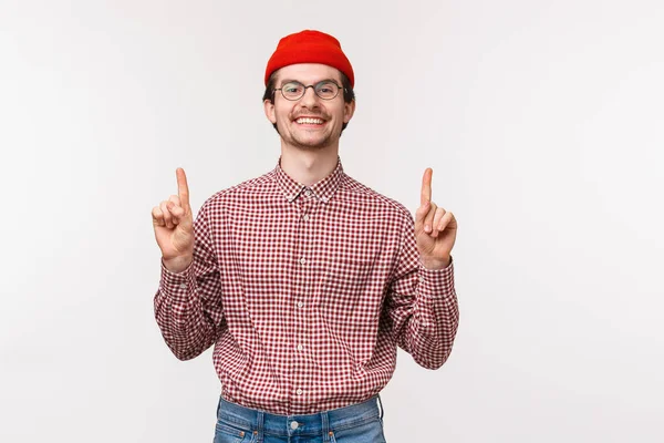 Waist-up πορτρέτο χαρούμενος περήφανος χαμογελαστός άνθρωπος σε γυαλιά και κόκκινο σκούφο, την προώθηση των προϊόντων ή της εταιρείας πανό, κοίτα κάμερα ως συμβουλή check out promo στην κορυφή, δείχνοντας προς τα πάνω, σταθεί λευκό φόντο — Φωτογραφία Αρχείου