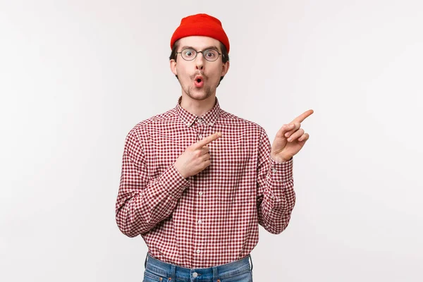 Έκπληκτος και περίεργος αστείο γενειοφόρος άνθρωπος σε κόκκινο σκούφο, γυαλιά πτυσσόμενα χείλη από έκπληξη και ενδιαφέρον ερώτηση σχετικά με δροσερό νέο προϊόν σε απόθεμα, δείχνοντας πάνω δεξιά γωνία — Φωτογραφία Αρχείου