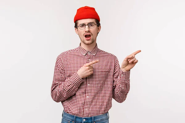 Portrait taille haute peu impressionné jeune homme réticent et ennuyé dans des lunettes et bonnet rouge bâillant désintéressé comme pointant coin supérieur droit à quelque chose qui ne vaut pas la peine d'attention, fond blanc — Photo