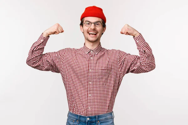 Midja-up porträtt av uppmuntrad och energisk ung säker smal kille i röd mössa och glasögon flex biceps, visar hans muskler är stark och kraftfull, stående vit bakgrund — Stockfoto