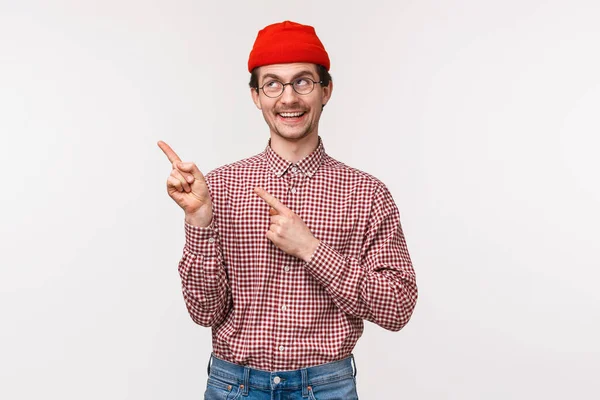 Portrait taille haute du gars geek souriant heureux dans des lunettes et bonnet rouge, pointant et regardant coin supérieur gauche avec sourire heureux, satisfait trouvé bon produit, recommander utiliser ce service — Photo