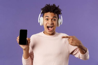 Teknoloji ve yaşam tarzı konsepti. Heyecanlı genç adamın portresi kulaklıklı müzik dinleyen, çevrimiçi platformda eğlenen ya da cep telefonuna işaret eden,