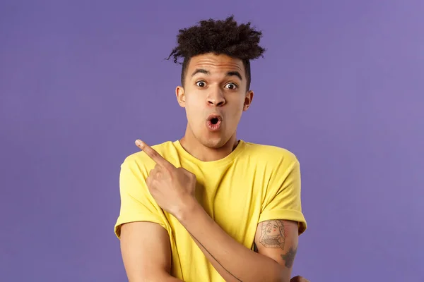 Portrait of surprised, excited young man showing cool new thing, pointing upper left corner, gasping, folding lips, stare camera impressed say wow, standing purple background — Stock Photo, Image
