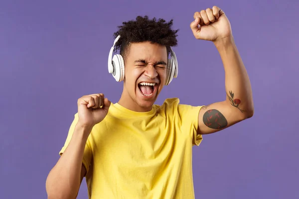 Primer plano retrato de alegre, joven bailarín feliz levantar la mano cantando, cerrar los ojos y sonriendo optimista como escuchar canción impresionante en los auriculares, disfrutando de la música, fondo púrpura —  Fotos de Stock