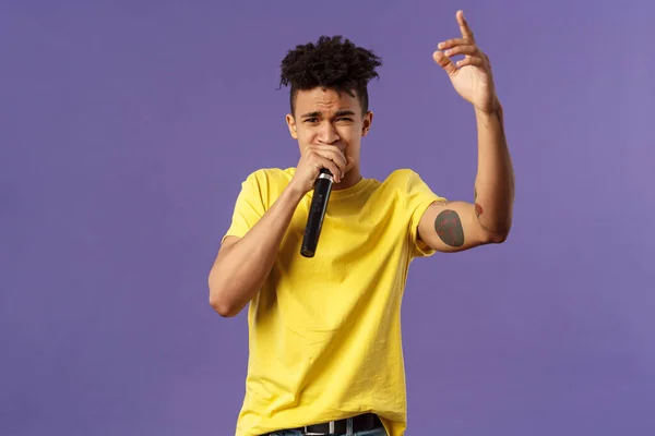 Porträt eines unbekümmerten jungen Hipster mit Tätowierungen, gelbem T-Shirt, der mit Hip-Hop oder Rap vor Publikum auftritt, Lieder singt, den Finger hebt, Mikrofon hält, Karaoke — Stockfoto
