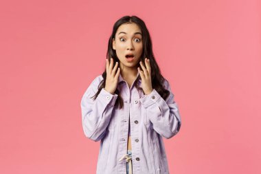 Şaşırmış Koreli genç bir kız şok edici bir şey gördü, kameraya şaşkınlıkla baktı, çenesini kapadı ve nefes nefese kaldı, büyük haberlerle el ele tutuşup, pembe arka planda durdu.