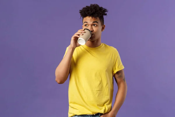 Tasasız genç bir erkek öğrencinin sabahları kafeinden enerji alması, kahve yudumlaması, ayakta dikilip bakması, kafede içki ısmarlaması, mor arka plan. — Stok fotoğraf